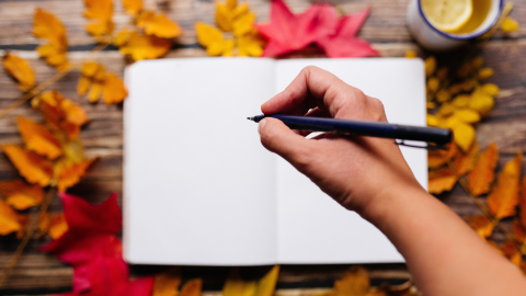 An open notebook on a table with fall leaves around it and a cup of tea with lemon and a hand hovering above the notebook with a pen.