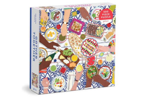 A jigsaw puzzle box with an image by Maja Tomljanovic titled, "Gather Together" and depicts a Mediterranean influenced vignette of friends and family gathering together at the table.