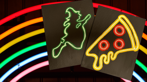 Neon rainbow on a black background with two paintings of with a neon outline of a witch flying, an a slice of pizza.